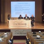 Escenario con mesa y cuatro ponentes, delante de una pantalla con una proyección